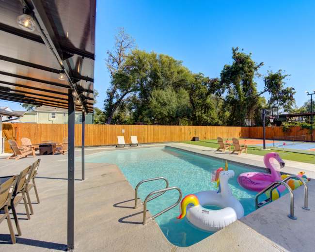 The image shows a swimming pool with colorful inflatable floats, surrounded by a wooden fence and adjacent to a basketball court.