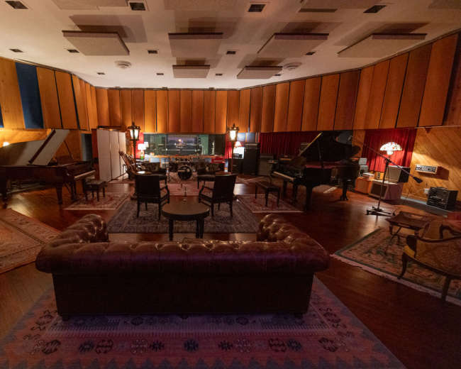 The image shows a vintage recording studio with multiple grand pianos, a mixing console in the background, and a cozy seating area featuring a leather sofa and several chairs.