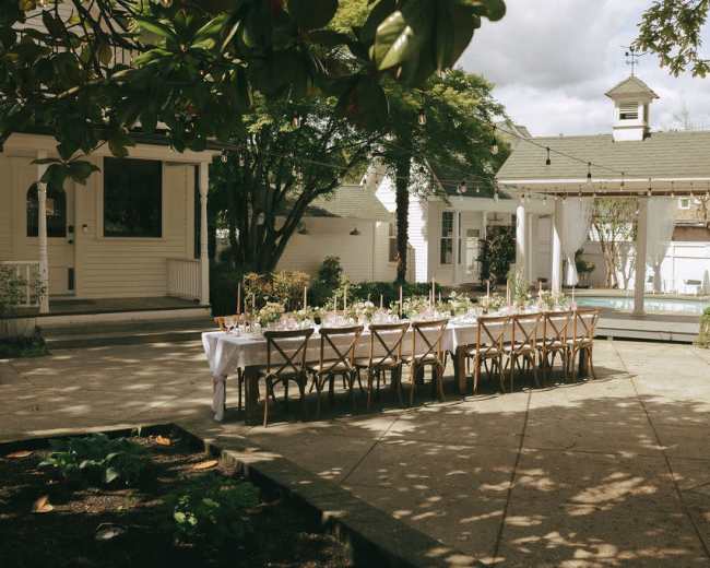 A long, elegantly set dining table is arranged outdoors beside a swimming pool, surrounded by greenery and a white gazebo.