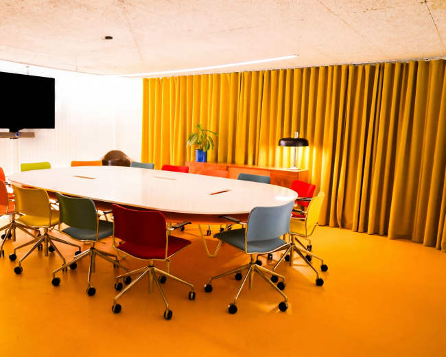 A bright and modern conference room features a large oval table surrounded by colorful chairs, with a television and large yellow curtains in the background.