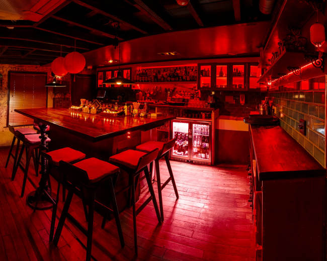 The image shows a dimly lit bar interior with a long wooden counter, high stools, and red ambient lighting.