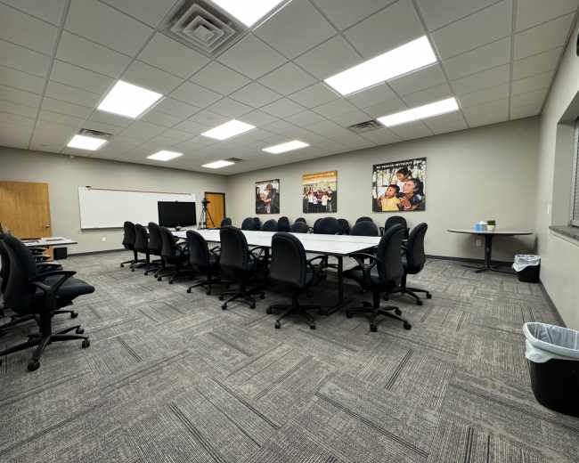 A large conference room features a rectangular table surrounded by black office chairs, with posters on the walls and a screen at one end.