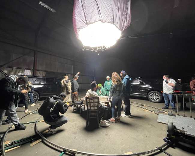 A group of people is gathered in a dimly lit warehouse, with film equipment and vehicles surrounding them during a shoot.