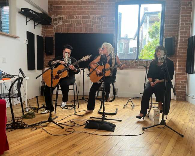 The image shows four musicians performing acoustically in a studio with brick walls and large windows letting in natural light.