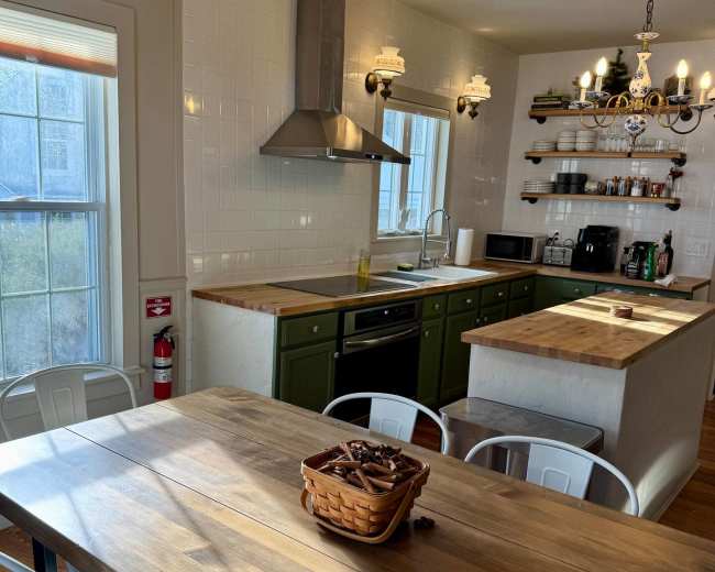 A kitchen features a wooden dining table with a basket in the center, modern appliances, and a decorative light fixture.