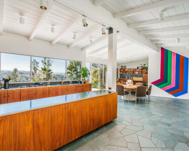 A modern kitchen with wooden cabinetry, a large island, and a dining area featuring a geometric artwork on the wall and large windows showing a view of greenery outside.