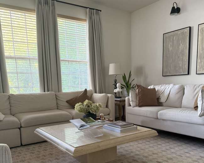 The image shows a cozy living room with two white sofas, a light-colored coffee table, and large windows adorned with curtains.