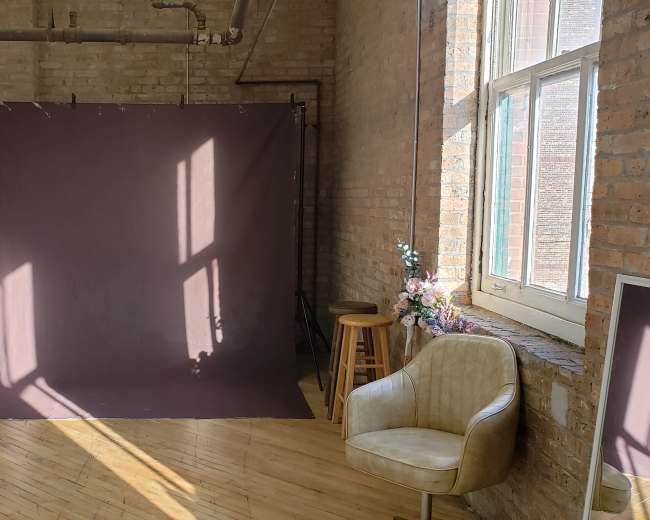 A spacious room features a brown backdrop, wooden flooring, a chair, and shelves with plants against a brick wall.
