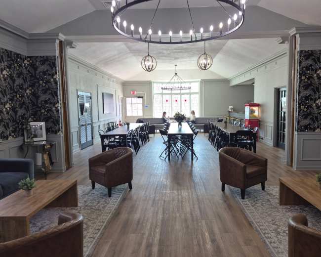 The image shows a spacious, elegantly decorated room with a long dining table surrounded by chairs, potted plants, and a seating area featuring brown leather chairs and tables.