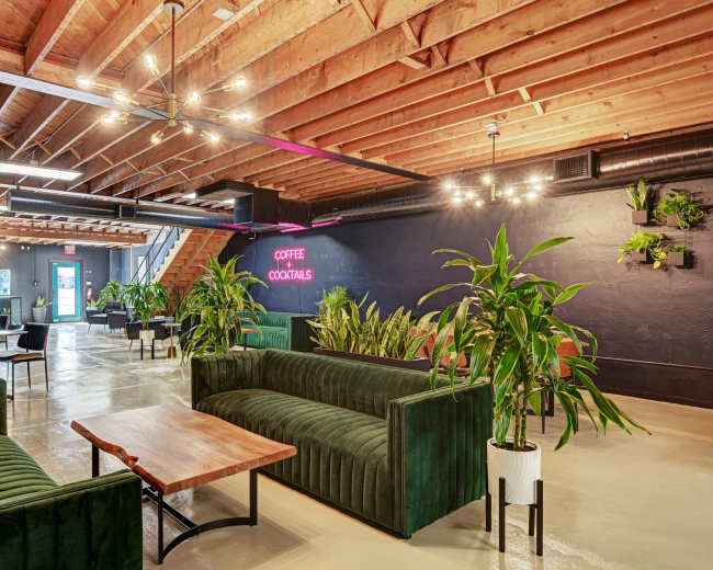 The image shows a modern lounge area featuring green velvet sofas, wooden furniture, and indoor plants against dark walls with a neon sign that reads "Coffee Cocktails."