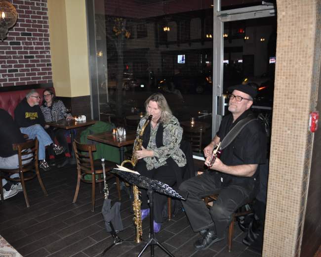 A saxophonist and a guitarist perform in a dimly lit restaurant, while patrons sit at tables nearby enjoying their meals.