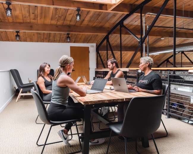 Four individuals are seated around a wooden table in a modern office space with exposed beams and large windows.