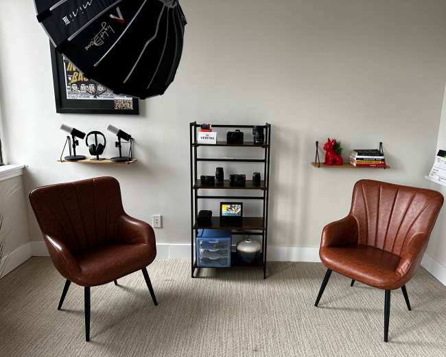 The image shows a minimalist room with two brown chairs facing a black shelving unit that holds cameras and books.