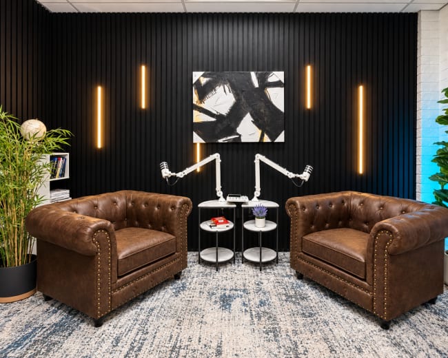 The image depicts a modern conversational area with two brown tufted leather armchairs, a circular side table between them, and a black wall adorned with vertical slats and recessed lighting.