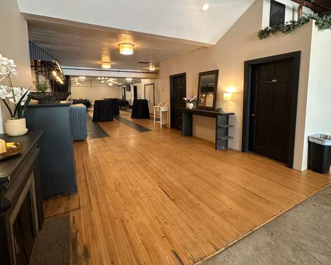 The image shows a spacious interior with wooden flooring, featuring a service area on the left, tables set in the background, and doorways on the right.