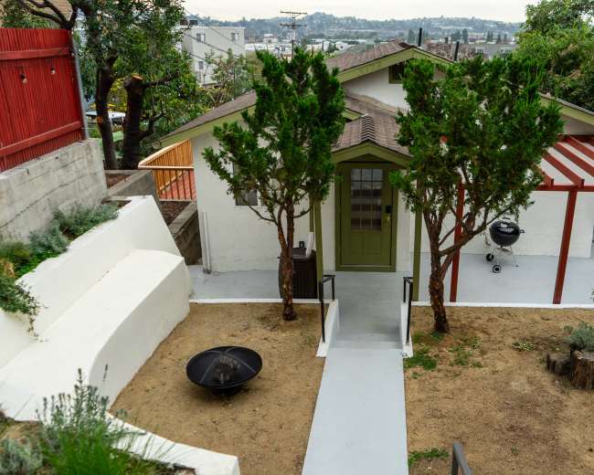 The image shows a house with a green door surrounded by two trees, a gravel area, and a small grilling station under a red pergola.