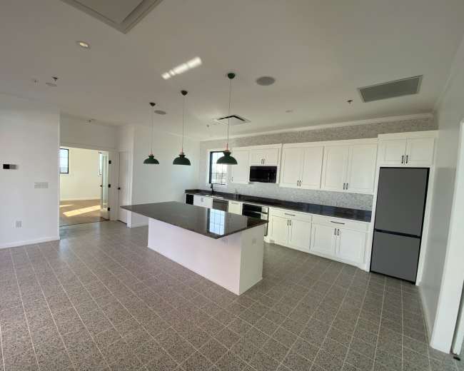 The image shows a modern kitchen with white cabinets, a dark countertop, and pendant lights, adjacent to a connected open space.