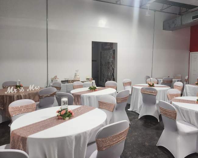 The image shows a banquet hall set up for a wedding reception, featuring round tables with white tablecloths and rose gold accents, along with a decorated dessert table in the background.