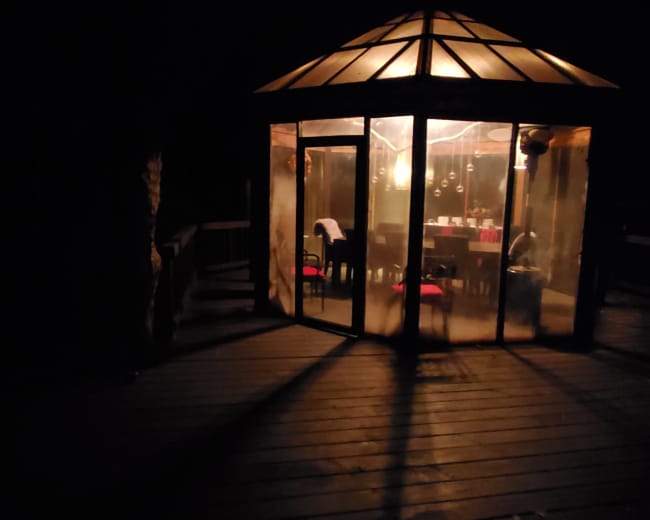 A glass-walled gazebo illuminated from within stands on a wooden deck at night.