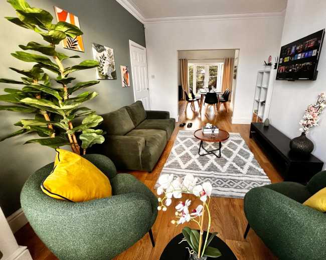The image shows a living room with a green sofa, two green chairs with yellow cushions, a round coffee table, and a decorative plant, leading to a dining area in the background.