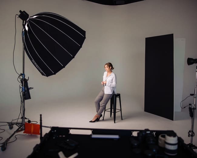 A woman sits on a stool in a photography studio, surrounded by lighting equipment and a black backdrop.