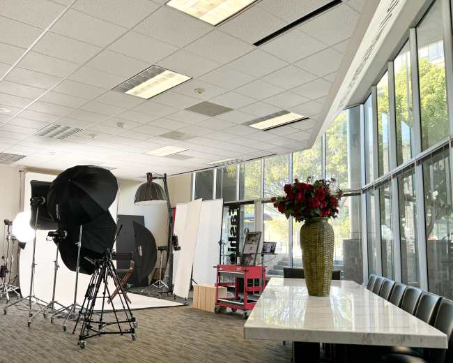 The image shows a photography studio with equipment and a large window, featuring a table with a vase of flowers.