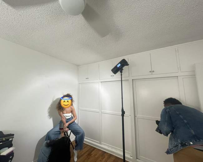 A person sits on a stool in a bare room while another person takes photos with a camera.