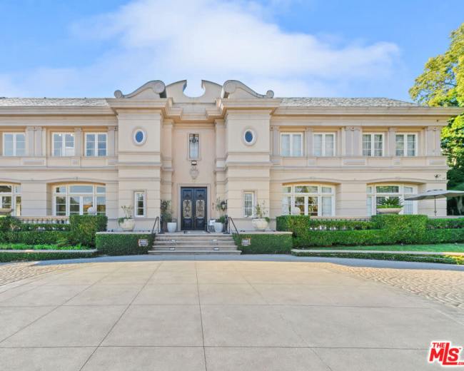 The image shows a large, stately mansion with a symmetrical façade, featuring a central entrance and landscaped gardens on either side.