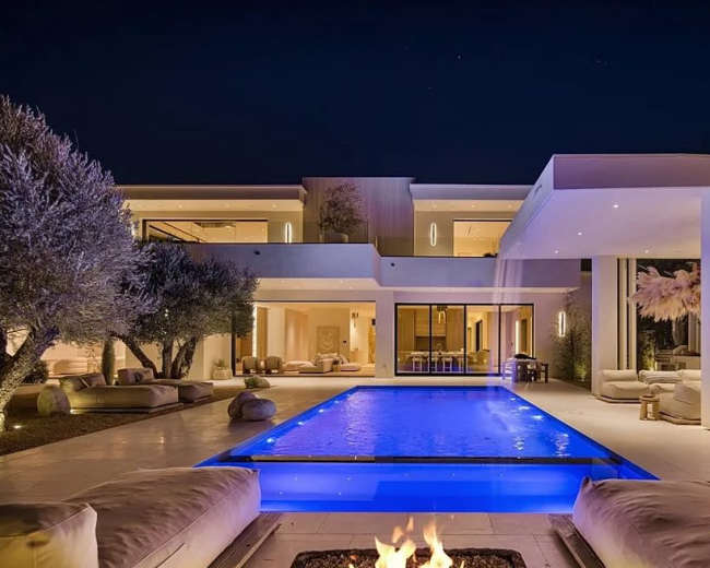 The image shows a modern villa at night with a brightly lit pool and a fire pit surrounded by lounge seating.