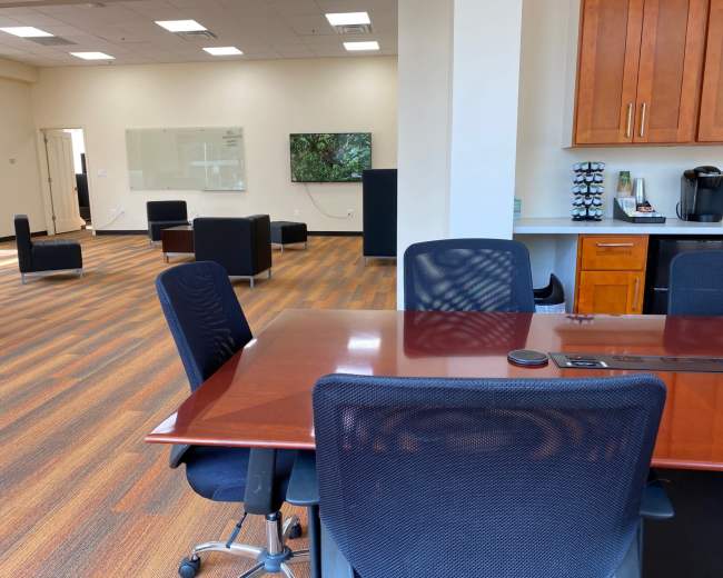 A modern office space featuring a conference table with chairs, a kitchen area with a coffee maker, and a lounge area with black seating.
