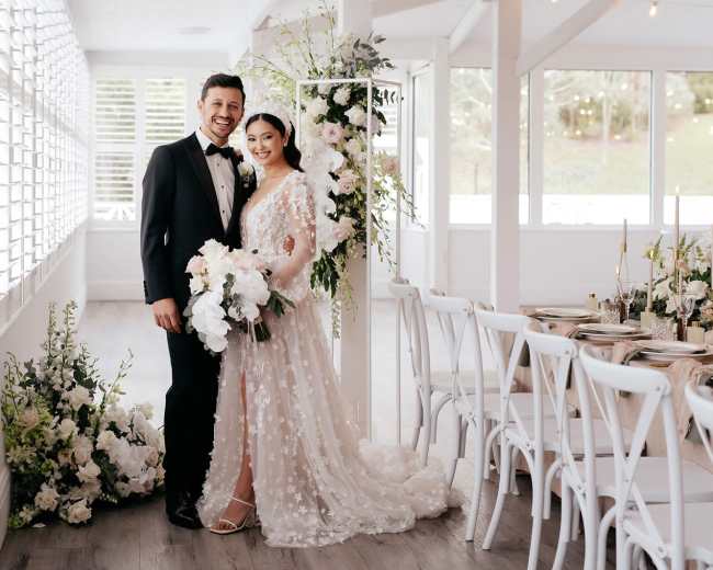 A couple stands together, smiling, in a bright venue decorated with flowers and elegant table settings for their wedding.