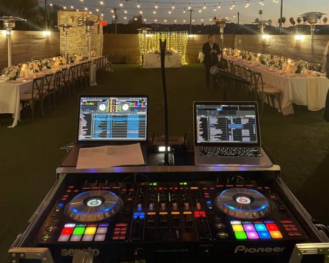 A DJ setup with turntables and laptops is positioned in front of an outdoor wedding reception featuring tables adorned with flowers and string lights, set against a twilight sky.