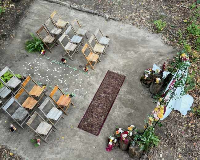 The image shows an outdoor wedding setup featuring wooden chairs arranged in rows on a dirt surface, with a decorative aisle runner and floral arrangements.