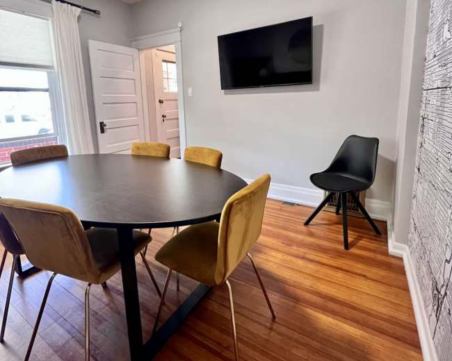 The image shows a dining area featuring a round black table surrounded by six yellow upholstered chairs, with a flat-screen TV mounted on the wall and a door leading to another room.