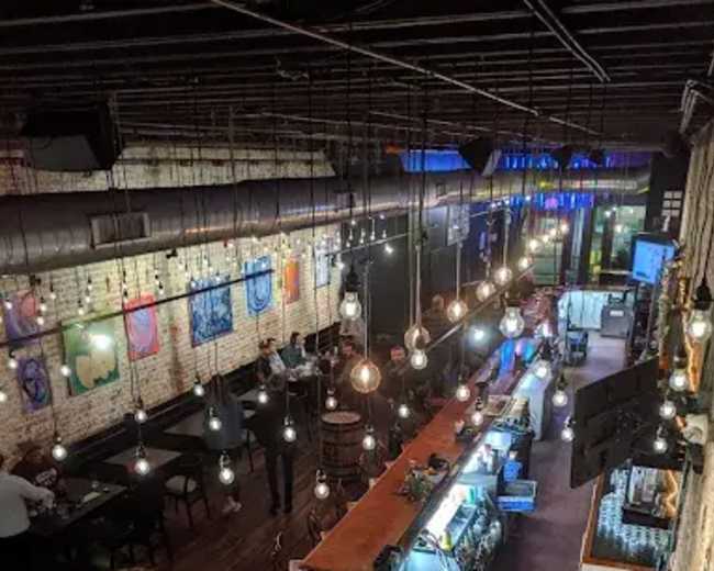 The image shows a dimly lit bar with exposed brick walls, wooden flooring, and hanging light bulbs, featuring a long counter and tables for seating.