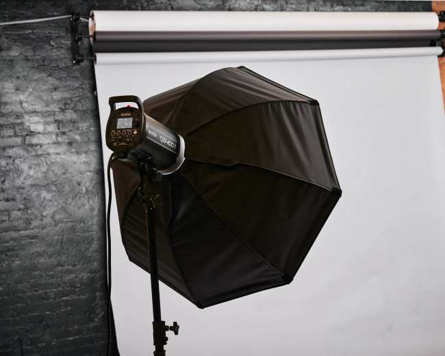A black umbrella softbox light is positioned in front of a white backdrop against a brick wall.