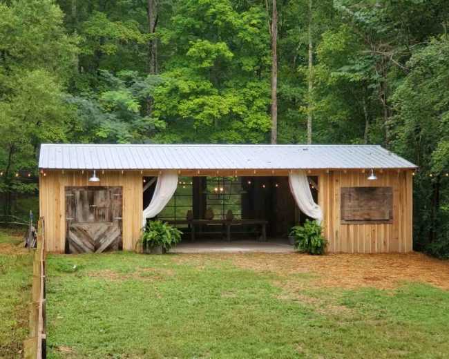A wooden cabin with a metal roof features open sides, decorative curtains, and is surrounded by greenery.