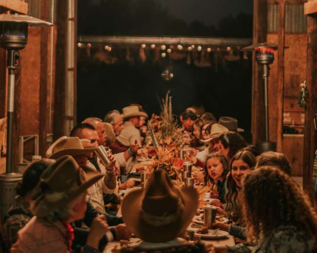 A long table set for a dinner gathering is illuminated by large circular chandeliers in a rustic barn at night.