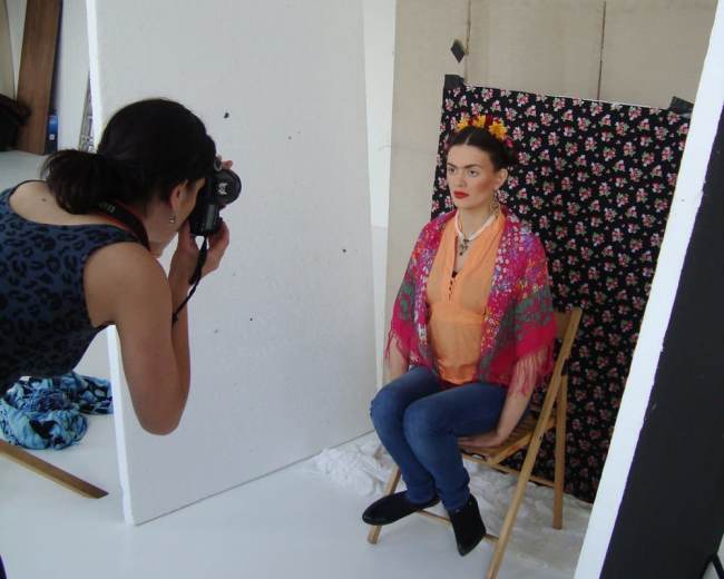 A woman is being photographed while sitting on a chair in front of a floral backdrop in a studio setting.