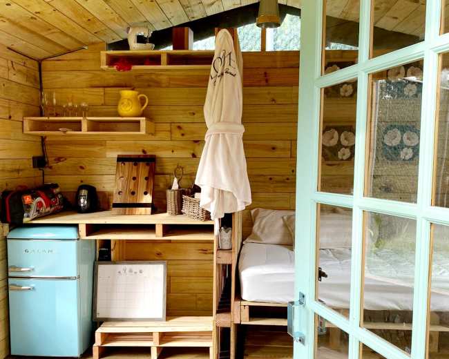 The image shows the interior of a small wooden cabin featuring a kitchenette, a bed area, and a door leading outside.