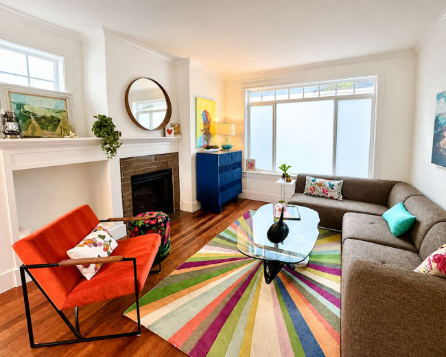 The image shows a modern living room featuring a colorful striped rug, a glass coffee table, a gray sofa, an orange chair, and a blue cabinet near a window.