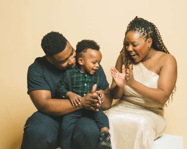 A family of three, seated on a white cube against a beige background, shares a joyful moment as they play and clap together.