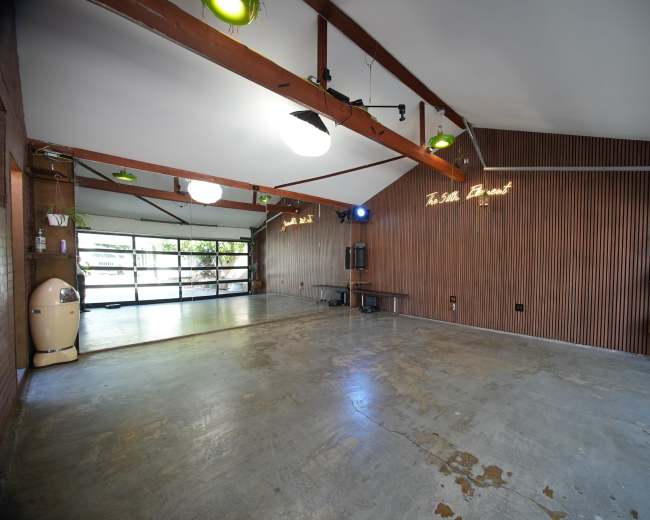 The image shows an empty room with wooden wall paneling, a concrete floor, and large glass doors that open to an outdoor area.