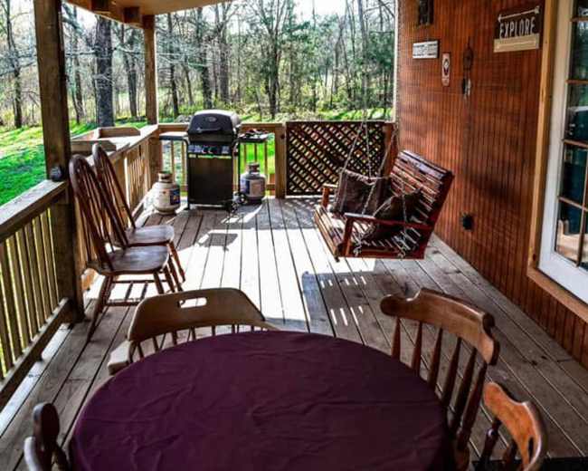 The image shows a wooden porch with a round table and four chairs, a rocking chair, a grill, and views of a forested area beyond.