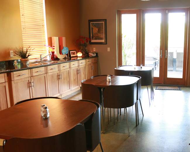 The image shows a small café with wooden tables, a countertop area, and large doors leading to an outdoor space.