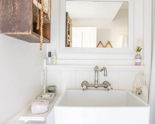 The image shows a clean bathroom with a wall-mounted mirror above a white sink and decorative elements on the shelves.