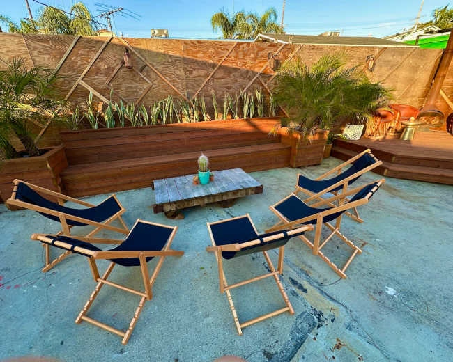 The image shows a seating arrangement of six folding chairs surrounding a low wooden table in an outdoor space with wooden planters and greenery.