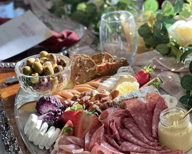A large platter of assorted meats, cheeses, fruits, crackers, and olives arranged artfully on a wooden table surrounded by greenery and tableware.