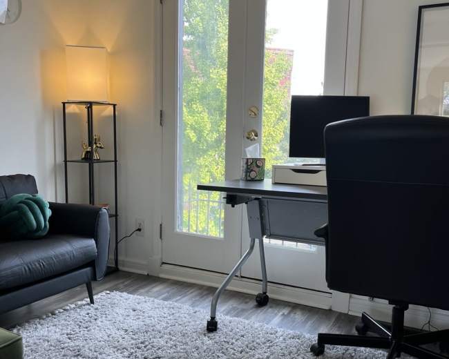 The image shows a cozy home office setup featuring a desk with a computer, a black office chair, a sofa, and a door leading to a balcony, all set on a plush rug.