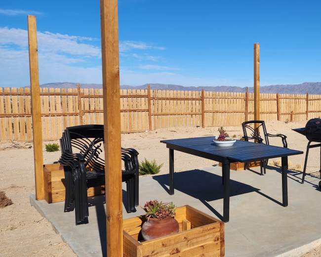 The image shows a simple outdoor seating area with a black table, two chairs, a grill, and potted plants, set in a sandy landscape with a wooden fence and mountains in the background.